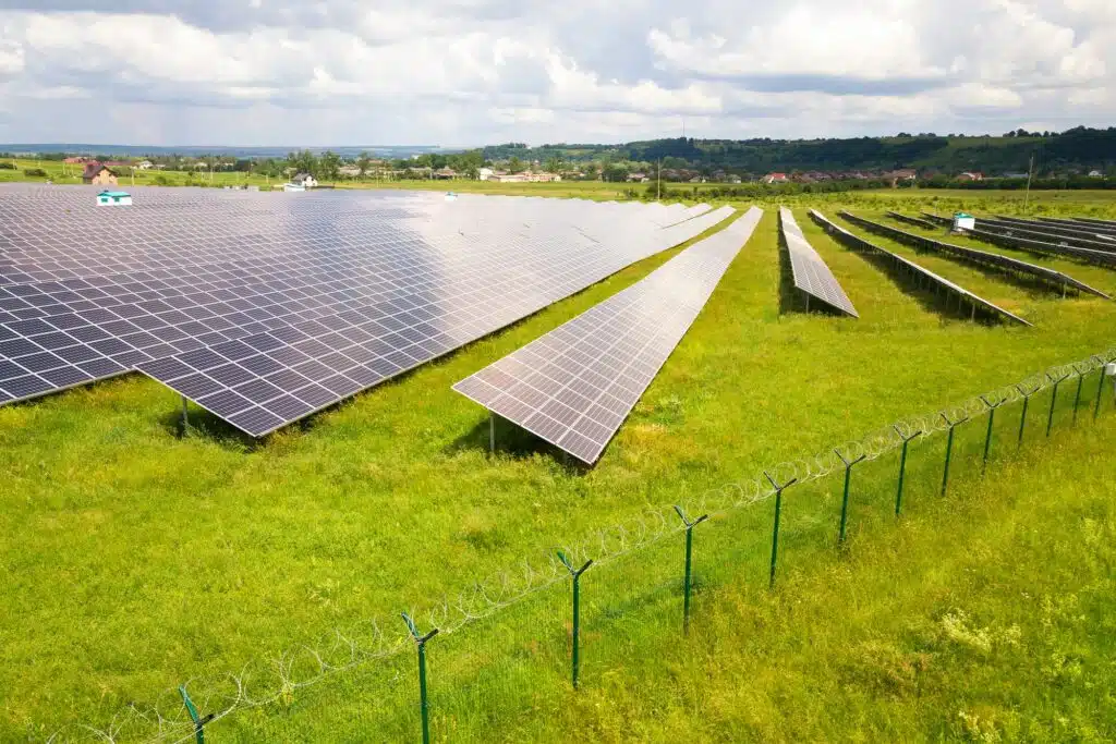 Luftaufnahme eines Solarkraftwerks auf einer grünen Wiese mit einem Maschendrahtzaun drum herum.
